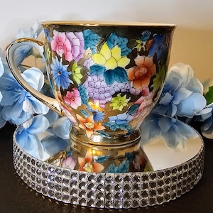 Könnte beinhalten: Eine dekorative Teetasse mit goldenem Henkel und Rand, verziert mit einem lebendigen Blumenmuster in Rosa, Blau, Gelb und Orange. Die Tasse steht auf einem Spiegeltablett mit einem funkelnden Kristallrand, das das farbenfrohe Design widerspiegelt.