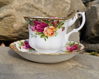 May include: White teacup and saucer set with a floral rose pattern in shades of pink, yellow, and green. The cup and saucer have gold trim. The teacup has a curved handle.