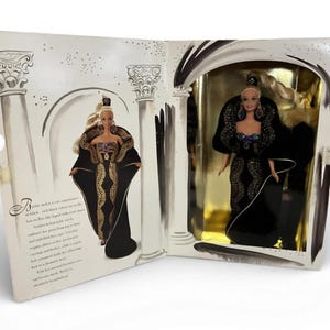 Puede incluir: Una muñeca Barbie con un vestido negro con detalles dorados y una capa a juego, presentada en su embalaje original. El embalaje presenta ilustraciones de columnas y arcos, con texto a la izquierda.