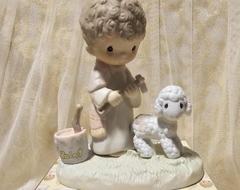 Może przedstawiać: Porcelanowa figurka dziecka z kręconymi włosami w szacie, trzymającego młotek i stojącego obok małego jagnięcia. Wiaderko z farbą z napisem Paint na boku stoi na trawiastej podstawie. Figurka posiada matowe wykończenie powierzchni.