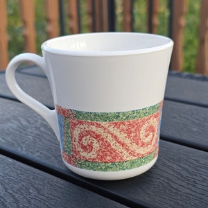Puede incluir: Taza de cerámica blanca con asa curvada. La taza presenta una banda decorativa con diseños rojos, verdes y azules. La taza está sobre una superficie oscura.