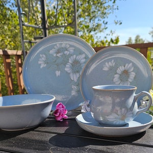 Puede incluir: Un juego de vajilla de cerámica azul claro con un diseño floral. El juego incluye un cuenco, platos, una taza y un platillo. Los platos y la taza presentan un patrón de margaritas blancas. El juego se exhibe sobre una mesa negra.