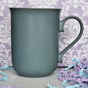 May include: A cylindrical mug with a matte, speckled teal-green finish. The mug features a curved handle on the right side and a slightly flared rim at the top. The surface has a uniform, granular texture throughout the exterior body.
