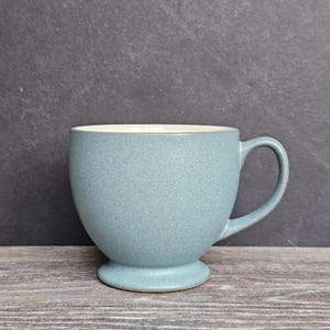 May include: A light blue ceramic mug with a speckled finish, featuring a rounded bowl-shaped body, a pedestal base, and a curved handle. The interior of the mug is a smooth, off-white color, contrasting with the textured blue exterior surface.