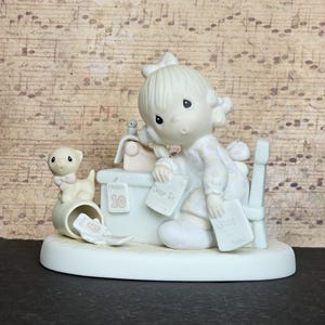 Könnte beinhalten: Eine Porzellanfigur eines Kindes, das an einem Schreibtisch mit einem kleinen Hund sitzt. Das Kind hält Papiere mit der Aufschrift Dear Sir und Bills. Ein Kalender auf dem Schreibtisch zeigt Friday 10. Der Hund sitzt neben einem Behälter mit Mon 6.
