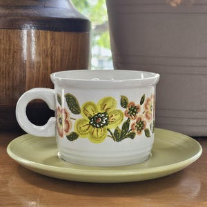 May include: A white ceramic coffee mug with a floral pattern in yellow, orange, and green, on a matching saucer. The mug has a rounded handle and a flared rim. The saucer and mug are on a wooden surface.