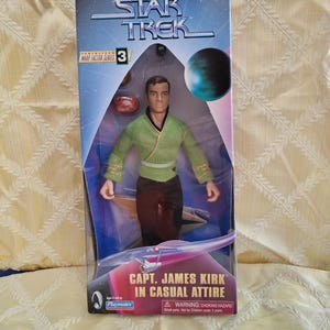 Może przedstawiać: Figurka akcji Star Trek Kapitan James Kirk w przezroczystym plastikowym opakowaniu. Figurka ma na sobie zieloną bluzkę kopertową, czarne spodnie i złote akcenty. Na opakowaniu napis "CAPT. JAMES KIRK IN CASUAL ATTIRE."