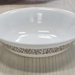 Puede incluir: Cuenco de cerámica blanco con un patrón floral marrón alrededor del borde. El cuenco tiene un acabado liso y brillante y está sobre una superficie de madera clara. Un clásico para servir alimentos.