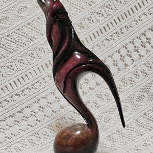 May include: A stylized sculpture of a howling wolf in shades of burgundy and black. The wolf is depicted with its head raised, mouth open, and body in a curved pose. The sculpture sits on a round base and is made of a glossy material.