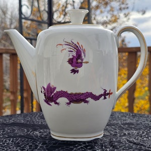 May include: White porcelain teapot with a lid, decorated with a purple dragon and bird motif. The teapot has a gold trim and a curved handle. The background is blurred, showing a wooden deck and foliage.