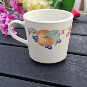 May include: Cream-colored ceramic mug with a curved handle. The mug features a decorative design of an orange fruit, blueberries, and red berries with green leaves. The mug is set against a dark wooden surface.