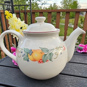 Könnte beinhalten: Cremefarbene Keramik-Teekanne mit Deckel und gebogenem Ausguss. Die Teekanne hat ein dekoratives Band aus gemalten Früchten und Blättern. Der Rand des Deckels ist mit einer roten Linie verziert. Die Teekanne steht auf einer dunklen Oberfläche.