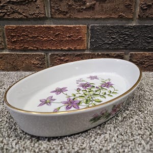 May include: An oval white porcelain serving dish featuring a delicate botanical illustration of purple flowers with green leaves on the interior base. The rim of the dish is finished with a thin gold metallic band, adding a refined detail.