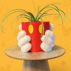 Puede incluir: Una maceta roja con círculos amarillos, que se asemeja a Mickey Mouse, contiene una planta verde. Unas manos blancas enguantadas parecen sujetar la maceta. La maceta está sobre un soporte de madera sobre un fondo amarillo con siluetas de Mickey Mouse.