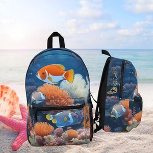 May include: A backpack featuring a vibrant underwater scene with orange, white, and blue fish swimming among coral. The backpack has a black handle and zipper, with a side view showing the same design. The background is a beach with a starfish and seashell.
