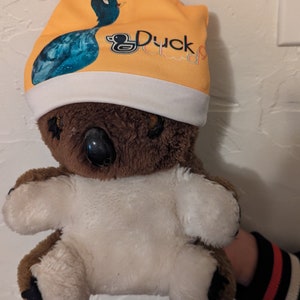Puede incluir: Un koala de peluche marr&oacute;n y blanco que lleva una boina amarilla y blanca con un pato azul y el texto "Duck Club" en ella.