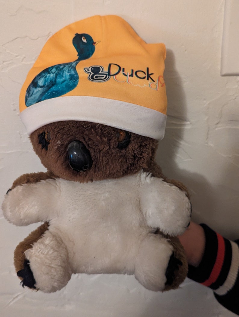 Puede incluir: Un koala de peluche marr&oacute;n y blanco que lleva una boina amarilla y blanca con un pato azul y el texto "Duck Club" en ella.
