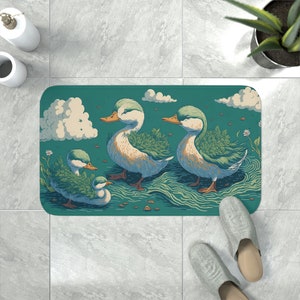 Könnte beinhalten: Eine tealblaue Badematte mit einem verspielten Design aus drei Enten mit grünem Laub auf ihren Köpfen. Die Enten schwimmen in einem blauen und grünen Wassermuster mit weißen Wolken im Hintergrund.