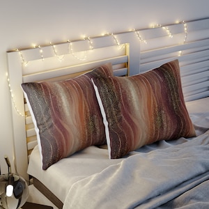 Peut inclure: Deux oreillers décoratifs avec un motif abstrait brun, or et blanc. Les oreillers sont sur un lit avec une tête de lit blanche et des guirlandes lumineuses.