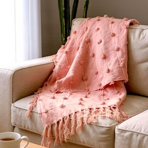 Puede incluir: Una manta rosa suave con un diseño texturizado y flecos está sobre un sofá beige. La manta presenta filas de detalles redondos en relieve y flecos en el borde inferior. Una taza de té y gafas están sobre una mesa de madera.