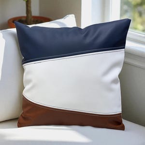Peut inclure: Un coussin décoratif avec un design en blocs de couleurs. La partie supérieure est bleu marine, le milieu est blanc et le bas est marron. Des coutures blanches accentuent les coutures. Le coussin est carré et repose sur un canapé blanc.