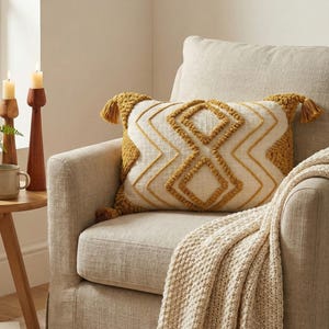 May include: A decorative rectangular pillow with a cream-colored background and a geometric pattern in mustard yellow. The pillow has matching yellow tassels on each end and is placed on a beige armchair. A cream-colored knitted throw is draped over the arm.