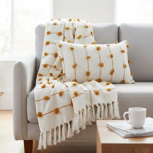 Op de afbeelding: Een witte plaid en kussenset met mosterdgele accenten ligt op een grijze bank. De plaid en het kussen hebben een textuurontwerp met kwastjes en pompons, wat een gezellige en uitnodigende uitstraling creëert.