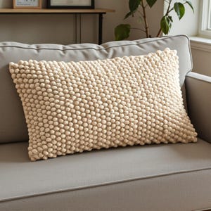May include: A rectangular beige decorative pillow with a textured, bobble-like surface. The pillow is placed on a gray sofa, offering a neutral color palette for home decor. The pillow's dimensions are not visible.