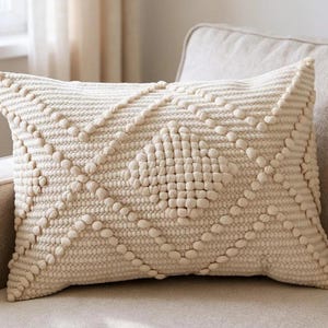 May include: Cream-coloured rectangular decorative cushion with a textured, raised pattern. The cushion features a geometric design of intersecting lines and a central diamond shape, all made with small, rounded bobbles. The cushion is placed on a neutral-toned sofa.