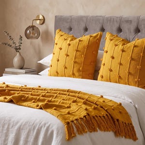 Könnte beinhalten: Ein Schlafzimmer mit einem Bett mit weißer Bettwäsche, zwei senfgelben Kissen und einer passenden Decke mit Fransen. Eine goldene Wandleuchte mit Glaskugel und eine Vase mit Grünzeug vervollständigen das gemütliche Ambiente.