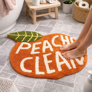 Puede incluir: Alfombra de baño con forma de melocotón naranja con las palabras "PEACH CLEAN" en blanco. La alfombra tiene un acento de hoja verde. La alfombra está sobre un suelo embaldosado.