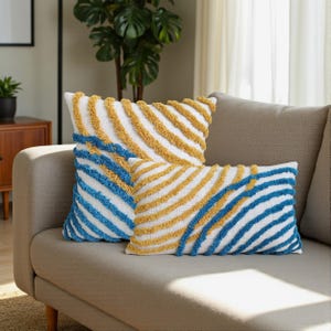 May include: Two decorative cushions with white fabric and textured, diagonal stripes in yellow and blue. The cushions are rectangular and square, resting on a beige sofa. The room has a neutral colour palette.