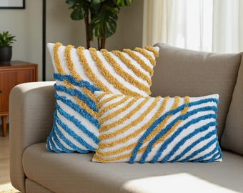 Könnte beinhalten: Zwei dekorative Kissen mit weißem Stoff und strukturierten, diagonalen Streifen in Gelb und Blau. Die Kissen sind rechteckig und quadratisch und liegen auf einem beigen Sofa. Der Raum hat eine neutrale Farbpalette.