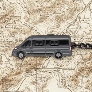 May include: A gray camper van keychain with a black chain and a large black ring. The keychain is set against a vintage map background, suggesting travel and adventure. The van has detailed windows and wheels.