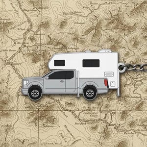 Puede incluir: Un llavero de camioneta camper plateada y blanca con una cadena y un llavero redondo. El llavero está sobre un fondo de mapa antiguo.