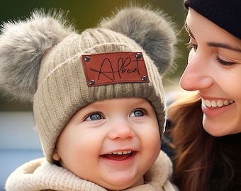 Könnte beinhalten: Ein lächelndes Baby trägt eine beige Strickmütze mit zwei grauen Bommeln und einem braunen Lederaufnäher mit der Aufschrift "Albert". Das Baby trägt einen cremefarbenen Pullover und wird von einer Frau mit schwarzer Mütze gehalten.