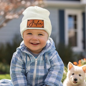 Peut inclure: Un bonnet crème avec un patch en cuir marron portant le nom "Julia" en cursive. Le bonnet est porté par un bébé souriant dans une combinaison à carreaux bleus. Un chiot blanc est assis sur l'herbe.