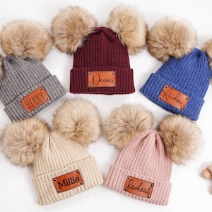 Puede incluir: Colección de seis gorros de punto acanalado en varios colores, cada uno con un pompón de piel sintética y una etiqueta con nombre de polipiel. Los colores incluyen gris, burdeos, azul, beige y rosa. Las etiquetas muestran nombres como "Herry", "Dennis" y "Andrew".