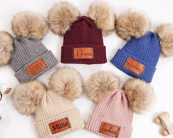 Könnte beinhalten: Sechs gerippte Strickmützen in verschiedenen Farben, jeweils mit einem Kunstfellbommel und einem Namensschild aus Kunstleder. Farben sind Grau, Burgunderrot, Blau, Beige und Rosa. Die Etiketten zeigen Namen wie "Herry", "Dennis" und "Andrew".