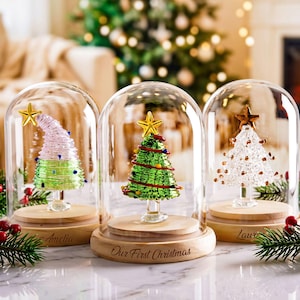 May include: Three glass Christmas tree ornaments enclosed in clear glass domes on light wood bases. The centre tree is green with a gold star, while the others are pink and white. The centre base is engraved with the text Our First Christmas.