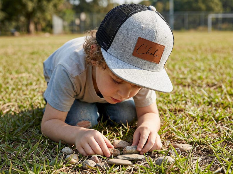May include: A gray and black trucker hat with a brown leather patch that reads "Chole" in a cursive font. The hat is worn by a person kneeling in a grassy field, examining small stones.