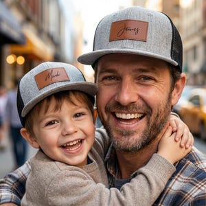 May include: Two gray and black trucker hats with brown leather patches. One hat has the name "James" and the other has "Micah" in script. The hats are worn by a smiling adult and child.