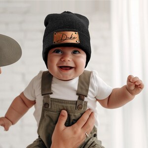 K&ouml;nnte beinhalten: Eine schwarze M&uuml;tze mit einem braunen Lederaufn&auml;her mit dem Namen "Duke" in Schreibschrift. Das Baby tr&auml;gt eine olivgr&uuml;ne Latzhose und ein wei&szlig;es Oberteil. Das Baby l&auml;chelt.