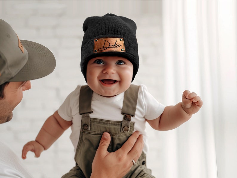 K&ouml;nnte beinhalten: Eine schwarze M&uuml;tze mit einem braunen Lederaufn&auml;her mit dem Namen "Duke" in Schreibschrift. Das Baby tr&auml;gt eine olivgr&uuml;ne Latzhose und ein wei&szlig;es Oberteil. Das Baby l&auml;chelt.