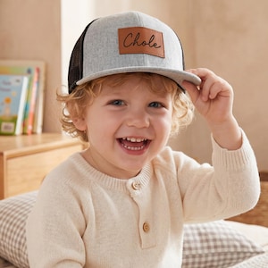 May include: A light gray and black trucker hat with the name "Chole" on a brown leather patch. The hat is worn by a smiling child with blond hair.