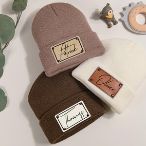 K&ouml;nnte beinhalten: Drei gestrickte M&uuml;tzen in Taupe, Wei&szlig; und Braun, jeweils mit einem rechteckigen Lederpatch. Die Patches sind mit den Namen "Patrick", "Oliver" und "Thomas" in Schreibschrift personalisiert. Ein Babyspielzeug und eine kleine B&auml;renfigur sind ebenfalls zu sehen.