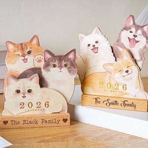 Pode incluir: Dois calendários de madeira de 2026 com ilustrações de gatos e cães. Um tem o texto "The Black Family" e o outro "The Smith Family". Os calendários estão sobre uma superfície branca.