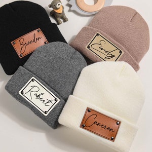 Pode incluir: Quatro gorros de malha em preto, cinza, rosa-púrpura e branco. Cada gorro tem um patch de couro marrom com um nome: "Brandon", "Robert", "Emily" e "Cameron". Os patches são fixados com pequenos rebites prateados. Os gorros são dobrados na borda.