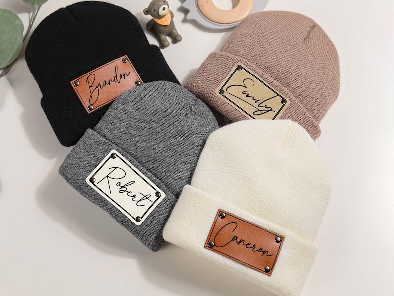 K&ouml;nnte beinhalten: Vier Strickm&uuml;tzen in Schwarz, Grau, Mauve und Wei&szlig;. Jede M&uuml;tze hat ein braunes Lederpatch mit einem Namen: "Brandon", "Robert", "Emily" und "Cameron". Die Patches sind mit kleinen silbernen Nieten befestigt. Die M&uuml;tzen sind am Rand umgeschlagen.