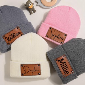 K&ouml;nnte beinhalten: Eine Auswahl an Baby-M&uuml;tzen in Grau, Rosa, Creme und Dunkelgrau. Jede M&uuml;tze hat ein braunes Lederpatch mit einem Namen in schwarzer Schrift. Die Namen sind Willian, Sophia, Daylan und Millie. Ein sch&ouml;nes Geschenk.